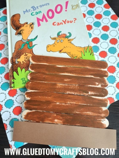 A Dr. Seuss book, “Mr. Brown Can Moo! Can You?”, sits on colorful patterned paper. In front, brown painted craft sticks are glued together with a brown paper strip—perfect for fun Dr Seuss Kid Crafts! Website “GLUEDTOMYCRAFTSBLOG.COM” is at the bottom.
