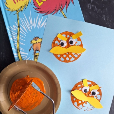 A copy of The Lorax book is next to a fun Dr. Seuss Crafts project with two Lorax faces made from orange paint, yellow paper eyebrows and mustaches, and big googly eyes on blue paper. A paint roller and plate are also visible.
