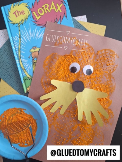 A Lorax-themed Dr. Seuss craft features an orange Lorax face made with bubble wrap print and yellow paper hand shapes for a mustache, with googly eyes, beside a copy of The Lorax book and crafting supplies.
