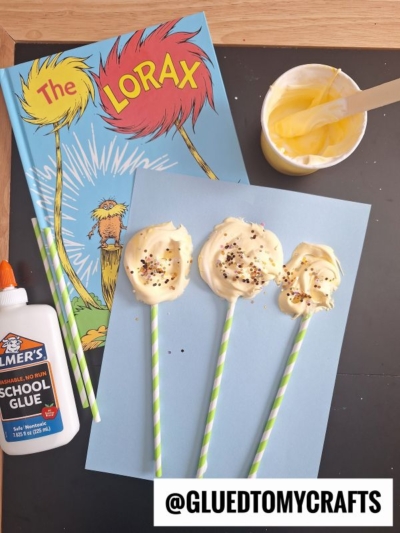 A flat lay of a Dr. Seuss The Lorax book, glue bottle, cup with yellow mixture and wooden stick, plus four paper straws with yellow blobs topped with sprinkles on blue paper—perfect inspiration for Dr Seuss Kid Crafts. @GLUEDTOMYCRAFTS text is at the bottom.
