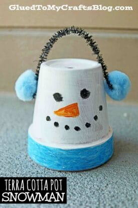 A cute terra cotta pot snowman craft, featuring a painted white face, black eyes and smile, an orange carrot nose, blue pom-pom earmuffs, and a blue band around the base. The small pot is displayed upside down for extra charm.