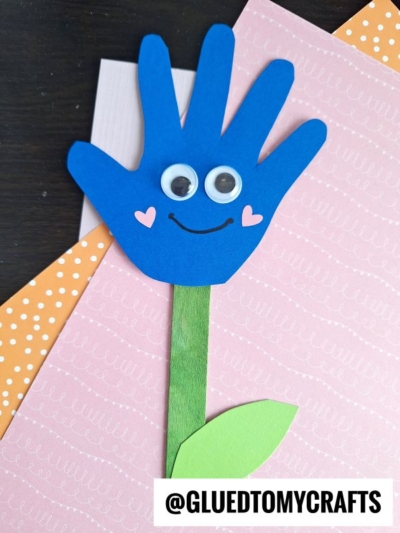 A blue handprint flower craft with googly eyes, pink heart cheeks, and a smile on pink and orange paper. A green stem and leaf are attached, making it a fun example of flowers made from handprints. @GLUEDTOMYCRAFTS is at the bottom.