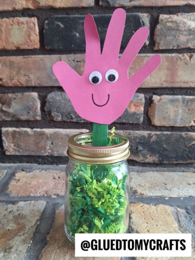 A craft project featuring a pink popsicle stick handprint flower with googly eyes and a smile, attached to a green stick in a jar filled with green shredded paper, with @GLUEDTOMYCRAFTS label at the bottom.