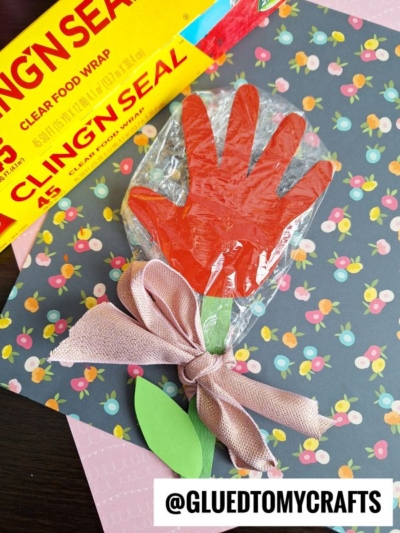 A handmade handprint flower craft made from a red paper handprint, green paper leaves, and pink ribbon, wrapped in clear plastic. The craft is displayed on a floral-patterned background with a Cling’n’Seal wrap box nearby.