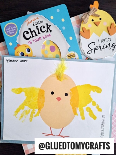 A handmade Easter card showcases a handprint easter chick with yellow wings, orange feet, and a feather on its head. It sits next to a children’s book about chicks and a wooden "Hello Spring" decoration.