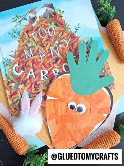 A children’s book titled “Too Many Carrots” sits on a table with a bunny ear headband, fake carrots, and a craft tissue paper carrot made from orange tissue paper, googly eyes, and a green handprint for leaves.
