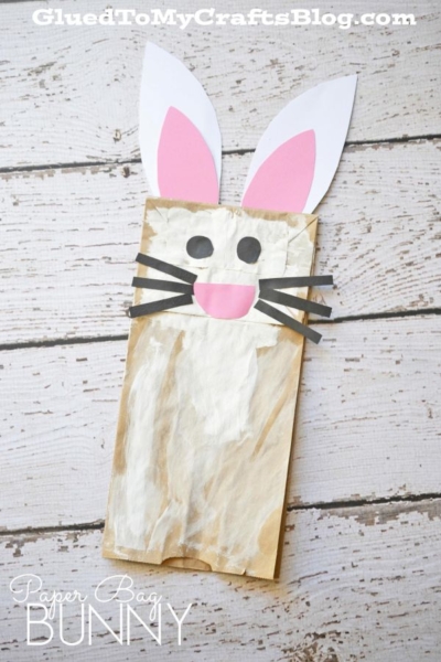 A paper bag bunny decorated with white paint, big paper ears, black whiskers, and oval eyes and nose rests on a wooden surface. Perfect for an Easter bunny paper bag craft. Text reads “Paper Bag Bunny.”.