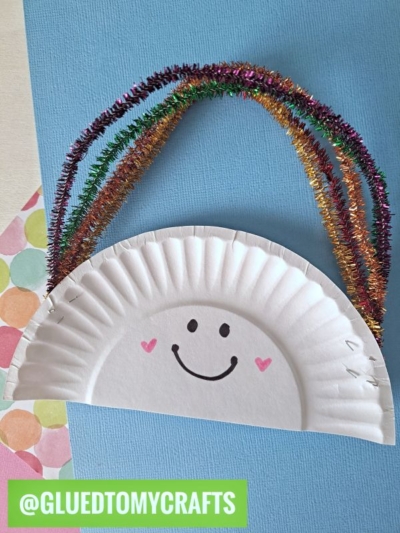 A paper plate folded in half with a drawn smiling face and pink cheeks. Glittery, colorful pipe cleaners form a rainbow on top—perfect for a St. Patty's Day Craft. A green label reads @GLUEDTOMYCRAFTS.