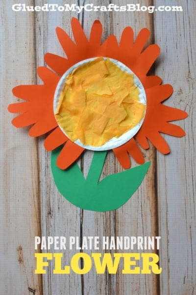 A paper plate handprint flower craft with vibrant orange handprint petals, a yellow tissue center, and green paper leaves and stem displayed on a wooden background. Text reads PAPER PLATE HANDPRINT FLOWER. This cheerful paper plate flower is perfect for kids’ crafting time!.