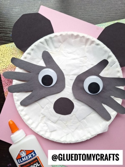 A cute paper plate panda features black paper ears, googly eyes, and cutout details for the nose and markings. This fun paper panda craft uses colored paper and supplies, which are visible in the background.