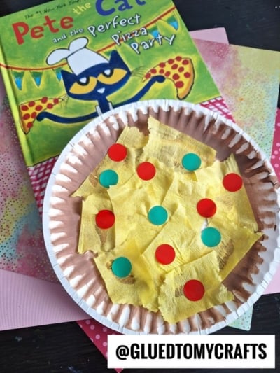 A children’s book, Pete the Cat and the Perfect Pizza Party, sits beside a colorful tissue paper pizza craft—a playful paper plate pepperoni pizza with yellow cheese and red and green toppings. The @GLUEDTOMYCRAFTS handle is visible.