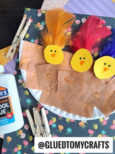 A kids’ craft project features three yellow paper chicks with feather crests in a paper plate bird nest, covered with brown paper squares, on a floral background. Clothespins and a glue bottle are nearby. @GLUEDTOMYCRAFTS is at the bottom.