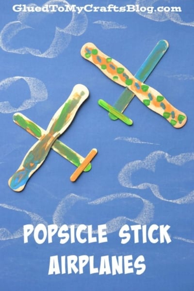 Two colorful, painted popsicle stick airplanes are displayed on a blue background with white chalk-drawn clouds. Text reads, POPSICLE STICK AIRPLANES—a fun popsicle stick airplane craft that shows you how to make an airplane out of popsicle sticks.