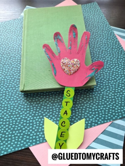 A handmade popsicle stick flower bookmark shaped like a red glittery hand with a heart rests on a green book. The stem spells "STACEY" with green circles and two yellow paper leaves. @GLUEDTOMYCRAFTS is at the bottom.