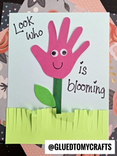 A pink popsicle stick flower with a handprint, googly eyes, and a smile sits on a card with green paper grass. The text says, “Look who is blooming.” The bottom features the tag @GLUEDTOMYCRAFTS.