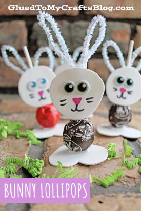 Three bunny lollipops with white pipe cleaner ears, googly eyes, and drawn-on faces stand upright with candy bases. Green paper grass decorates the surface. Text reads Bunny Lollipops.