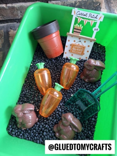 A carrot garden sensory bin filled with black beans, plastic carrots, toy rabbits, plant pots, scoops, and a Carrot Patch sign; a @GLUEDTOMYCRAFTS watermark appears at the bottom.