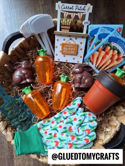 A basket contains gardening gloves with a carrot pattern, plastic carrot containers, chocolate bunny candies, seed packets, garden markers, flowerpots, a blue scoop, and a small āCarrot Patchā signāperfect for creating a carrot garden sensory bin. @GLUEDTOMYCRAFTS.