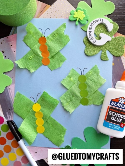 A St. Patty's Day Craft project featuring three green butterfly collages made from crepe paper and orange/yellow dot stickers on blue paper, surrounded by clover cutouts, glue, and a @GLUETOMYCRAFTS sticker.