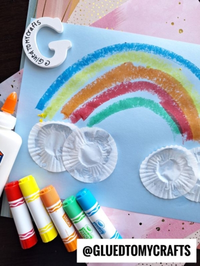 A paper craft shows a rainbow and clouds made from colored markers and cupcake liners on blue paper—a perfect St. Patty's Day craft. Nearby are four markers, a bottle of glue, and a white letter G with @GLUEDTOMYCRAFTS written on it.
