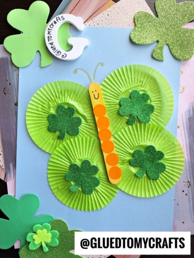 A St. Patty's Day craft butterfly made from green cupcake liners, glittery shamrocks, and an orange-dotted craft stick on blue paper. Shamrocks and a letter G accent the corners. @GLUEDTOMYCRAFTS label at the bottom.