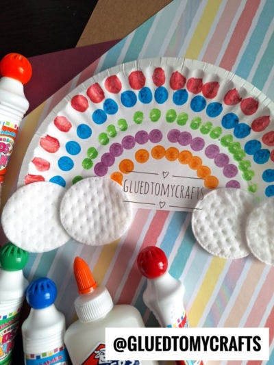 A St. Patty's Day Craft: A paper plate decorated with colorful paint dots forming a rainbow, cotton pads as clouds, glue bottles, and the text @GLUETOMYCRAFTS on a striped background.