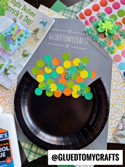 A St. Patty’s Day Craft features a black paper plate pot, topped with colorful paper circles and glittery shamrocks, surrounded by a book, glue, and patterned papers. Text reads @GLUEDTOMYCRAFTS.