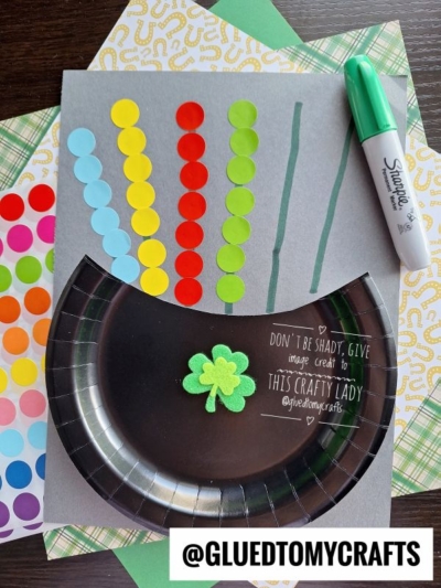 This St. Patty's Day Craft features a gray paper with green marker lines, colorful dot stickers as a rainbow, a paper plate adorned with a green shamrock, a green Sharpie, and patterned papers underneath.