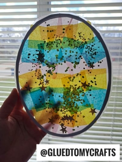 A hand holds up an oval-shaped stained glass Easter egg suncatcher, crafted with colorful tissue paper squares and star-shaped glitter, as sunlight shines through. The Instagram handle @GLUEDTOMYCRAFTS appears at the bottom.