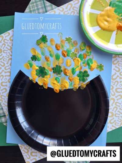 A blue paper with a black paper plate half glued on to resemble a pot. Above it, green and yellow paint fingerprints create clovers and coins for a fun St. Patty's Day Craft. Craft materials are visible in the background. Text reads “@GLUEDTOMYCRAFTS.”.
