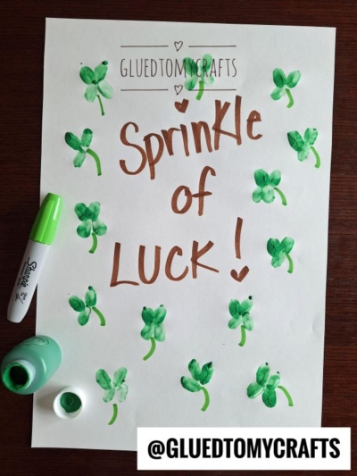A white paper with “Sprinkle of Luck!” written in brown, surrounded by green marker and paint shamrocks—perfect for a St. Patty's Day Craft. A green marker, green paint bottle, and the handle @GLUETOMYCRAFTS appear beside the paper.
