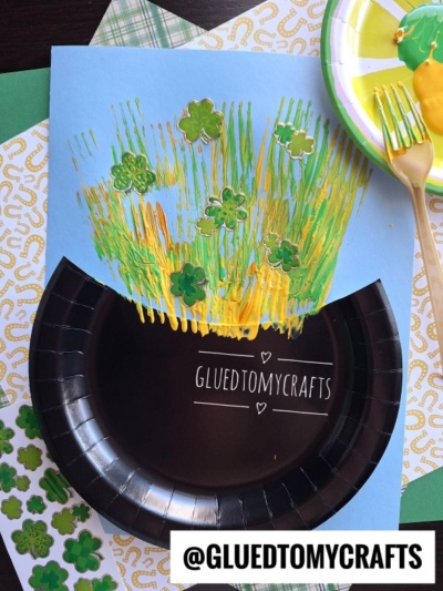 This St. Patty's Day Craft features a paper plate painted black as a pot, blue paper with yellow and green fork-painted lines for gold, and green shamrock stickers. Nearby are a plastic fork and paint palette.