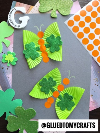 St. Patty's Day Craft featuring butterfly creations with green cupcake liner wings, glittery shamrocks, and orange dot stickers—arranged on a gray background with shamrock cutouts and craft supplies. @GLUEDTOMYCRAFTS is displayed at the bottom.