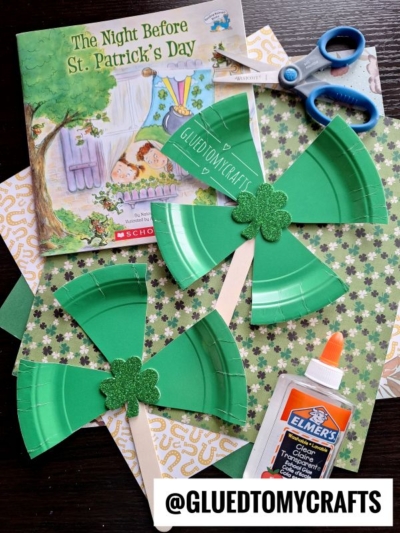 A St. Patty’s Day Craft scene featuring a book, green shamrock pinwheels made from paper plates, patterned paper, scissors, glue, and a popsicle stick. The @GLUEDTOMYCRAFTS label sits at the bottom for festive inspiration.