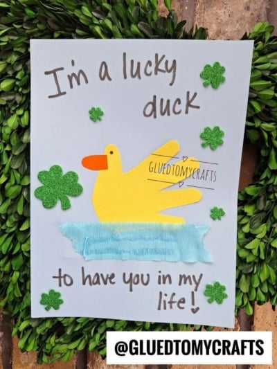 A handmade St. Patty's Day Craft card features a yellow duck, made from a handprint, swimming on blue water. Green shamrocks are scattered around, with the text “I’m a lucky duck to have you in my life!” @GLUEDTOMYCRAFTS is at the bottom.
