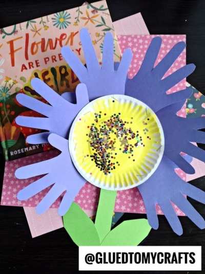 A colorful paper plate handprint flower craft made with purple handprint cutouts as petals, a yellow paper plate center with glitter, and green paper leaves, shown next to a book and pink patterned paper. @GLUEDTOMYCRAFTS is at the bottom.