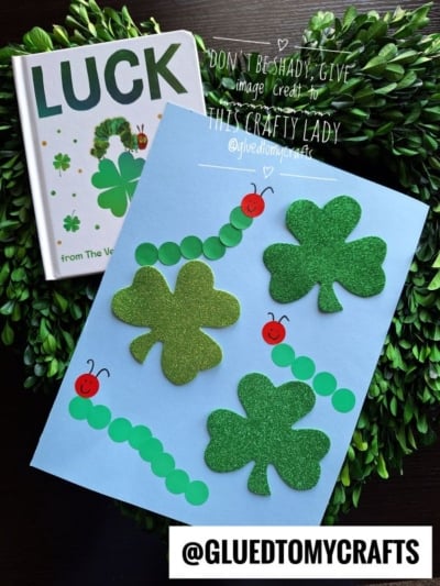 A blue paper showcases a St. Patty's Day Craft with three glittery green shamrocks and two caterpillars made from green dots and red heads. A LUCK book and greenery sit nearby. @GLUEDTOMYCRAFTS is labeled at the bottom.