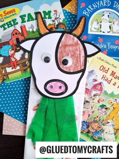 A paper bag cow puppet, also known as a lunch sack cow, with a big googly eye and green scarf sits on top of children’s farm-themed books, including Dr. Seuss’s The Farm, Barnyard Dance! and Old MacDonald Had a Farm.
