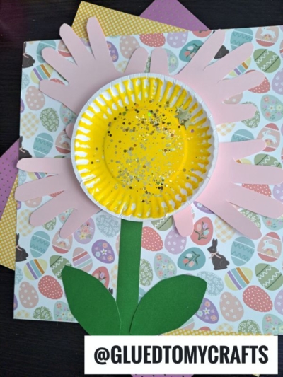 A handmade paper plate handprint flower craft with pink paper handprints as petals, a yellow cupcake liner center, and green leaves and stem, set on Easter-themed patterned paper. @GluedToMyCrafts is written at the bottom.