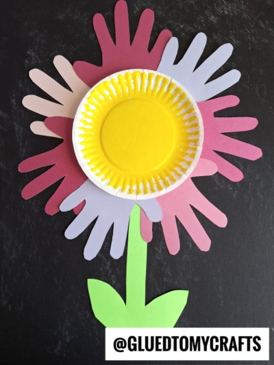 A paper plate handprint flower craft made from colorful handprint cutouts as petals, a yellow painted cupcake liner center, and green paper stem and leaves on a black background. @GLUEDTOMYCRAFTS is written at the bottom.