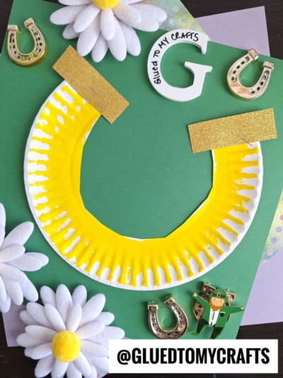 A St. Patty's Day craft featuring a paper plate painted yellow and cut into a horseshoe shape is displayed on green paper, surrounded by white daisies, gold horseshoe clips, and a “Glued to My Crafts” label.