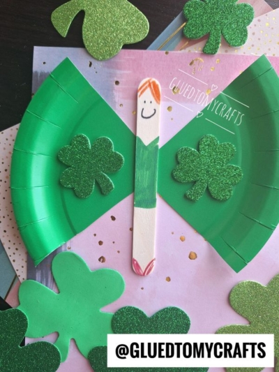A popsicle stick angel with a smiley face and green paper plate wings, decorated with sparkly green shamrocks, sits on a surface with other shamrock cutouts—a perfect St. Patty's Day Craft! @GLUEDTOMYCRAFTS is written at the bottom.