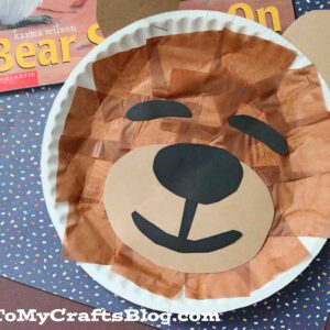 A paper plate sleeping bear craft, made with brown tissue paper, black paper nose and eyes, and paper ears, is displayed next to a children’s book