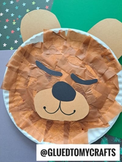 A paper plate sleeping bear craft made from a paper plate, brown tissue paper, and paper cutouts for facial features and ears, placed on colorful paper. Text @GLUEDTOMYCRAFTS is at the bottom.