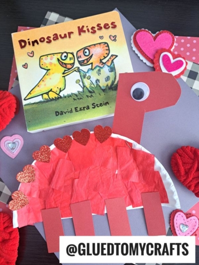 A children’s book titled Dinosaur Kisses is next to a red paper plate dinosaur craft made from paper and tissue, surrounded by heart decorations. A tag at the bottom reads @GLUEDTOMYCRAFTS.