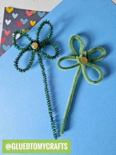 Two shamrock shapes made from green pipe cleaners with gold glitter centers sit on a blue surface. A heart-patterned paper is in the background, perfect for your next St. Patty's Day Craft. Label reads @GLUEDTOMYCRAFTS.
