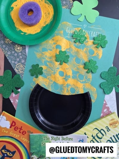A St. Patrick’s Day craft featuring a black paper plate as a pool noodle stamped pot of gold, glittery green shamrocks, and golden mesh spilling out. The background includes green, patterned papers and a label @GLUEDTOMYCRAFTS