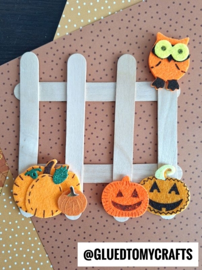 A Halloween craft popsicle stick picket fence, decorated with felt pumpkins and an owl, sits on polka dot paper. @GLUEDTOMYCRAFTS is written at the bottom.