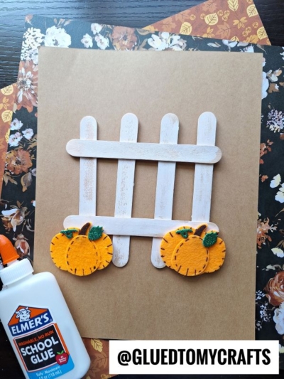 A mini popsicle stick picket fence, decorated with two felt pumpkins, is on a brown background with floral paper, a bottle of Elmers glue, and a @GLUEDTOMYCRAFTS label at the bottom.