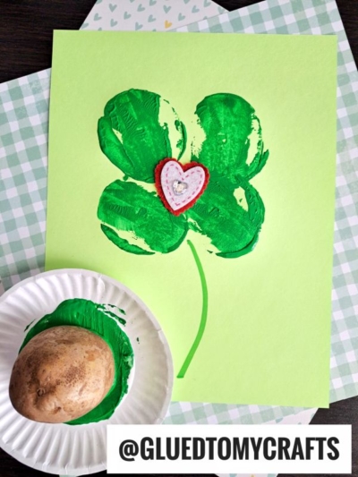 A St. Patty's Day craft featuring a shamrock stamped with green paint from a potato onto light green paper. A small red and white heart is glued in the center. The potato handle rests on a plate of green paint. @GLUEDTOMYCRAFTS is at the bottom.